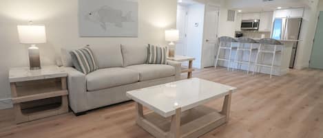 Uma sala de estar com sofá, mesa de centro e duas luminárias. É possível ver uma cozinha ao fundo, com geladeira e micro-ondas.