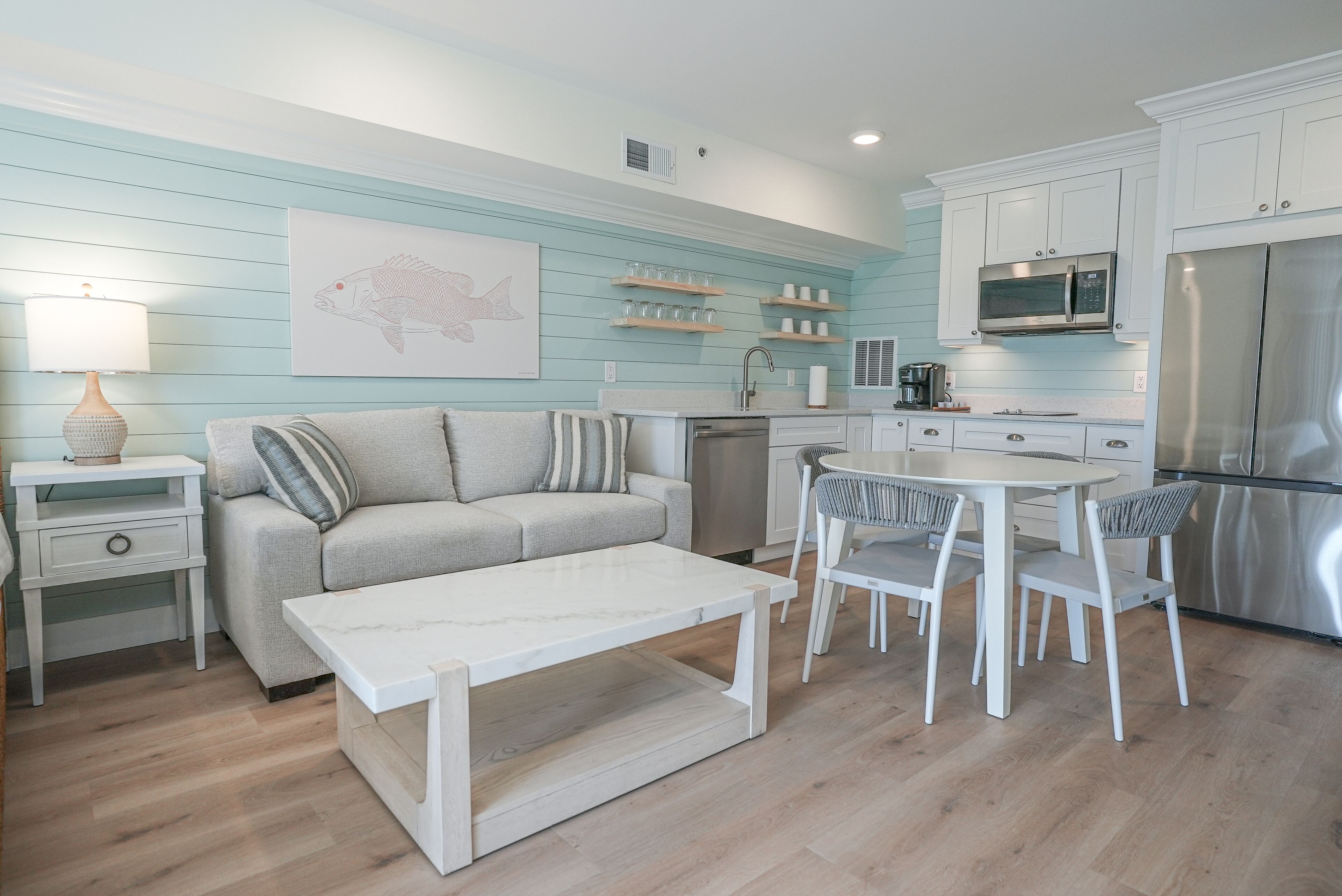 Una cocina moderna con armarios blancos, una zona de comedor con una mesa y sillas blancas, y una pared azul claro con un mural de peces.