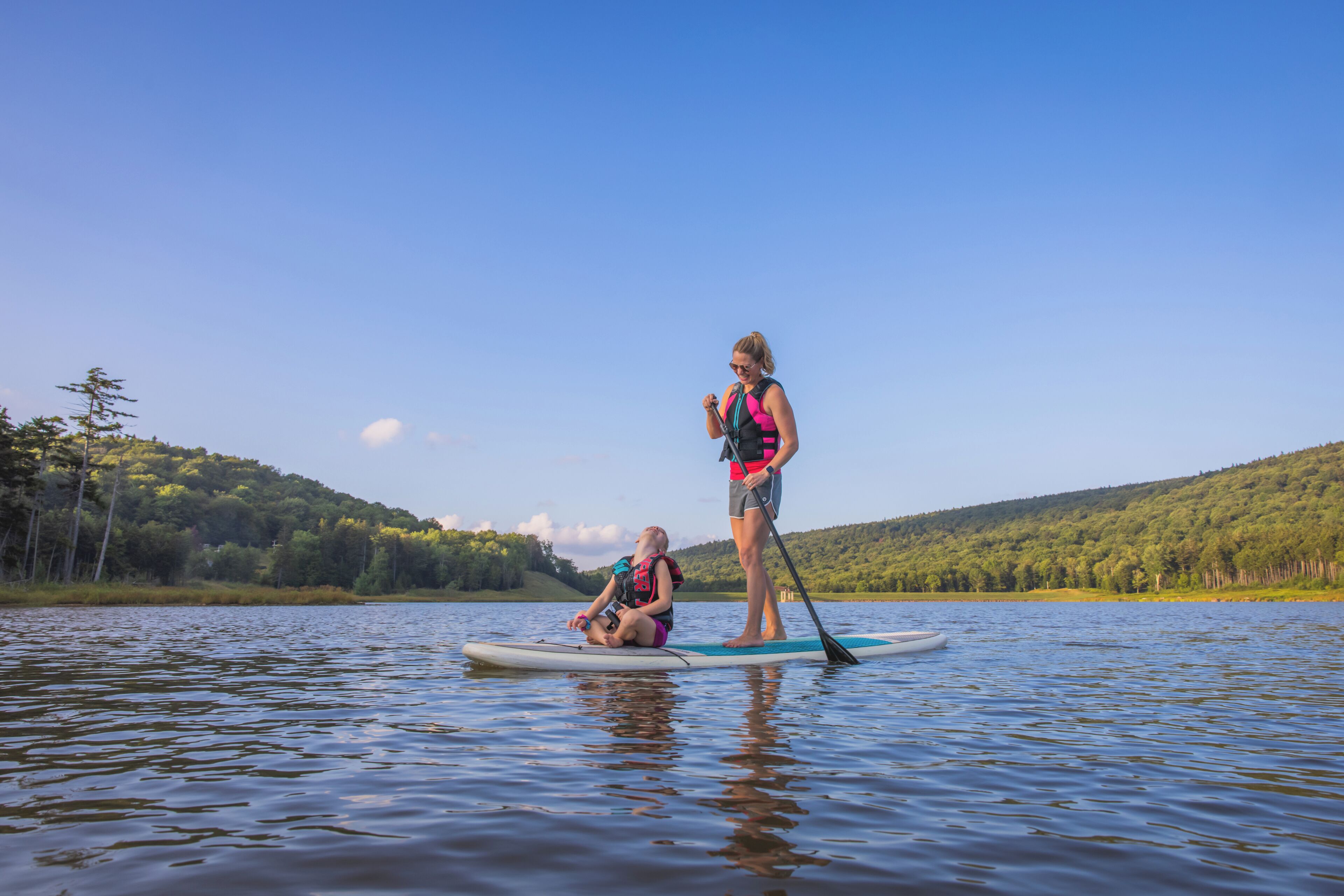 Two people on a paddleboard in a calm lake with a forested hill in the background.