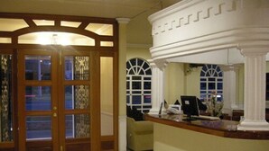A reception area with a wooden desk, a computer, and a telephone. There are two large windows with white frames and a decorative wooden partition with glass panels. - Scala Hotel (Lagos de Moreno)