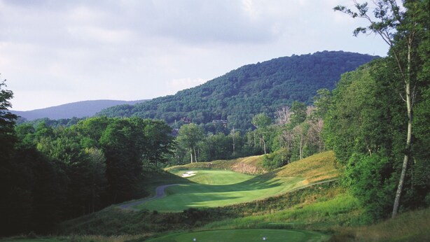 Un parcours de golf avec un sentier sinueux, un bunker de sable et une colline boisée en arrière-plan.