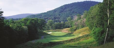 A golf course with a winding path, a sand bunker, and a forested hill in the background.