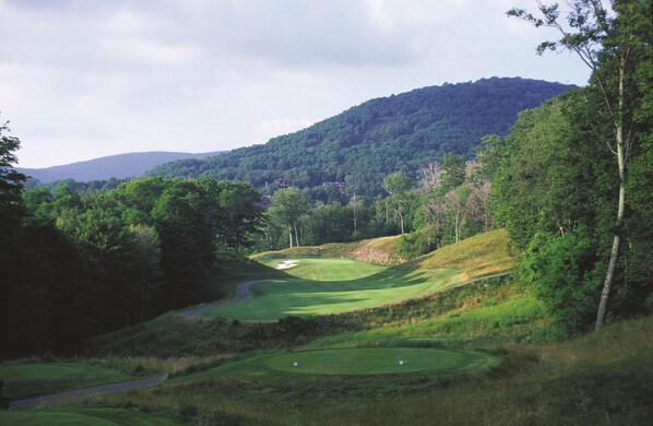 Un campo de golf con un sendero sinuoso, un búnker de arena y una colina boscosa al fondo.