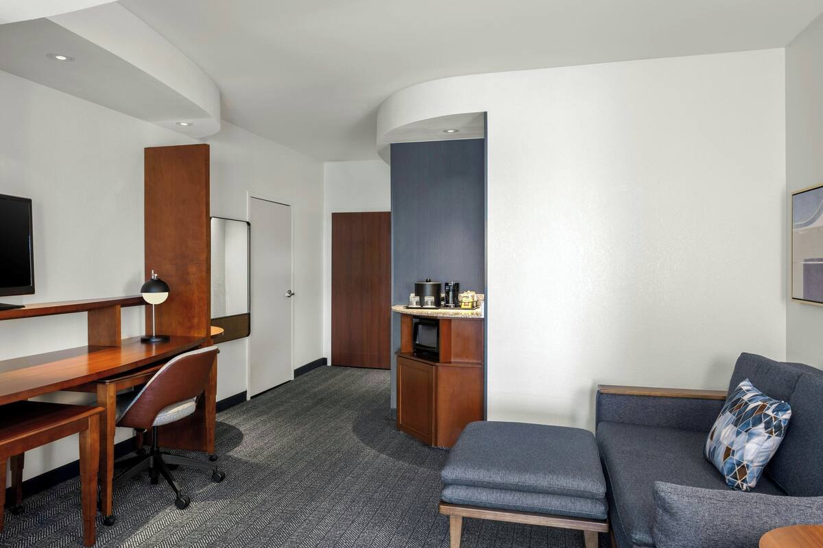 A hotel room with a wooden desk, a grey sofa, a TV, and a kitchenette area.