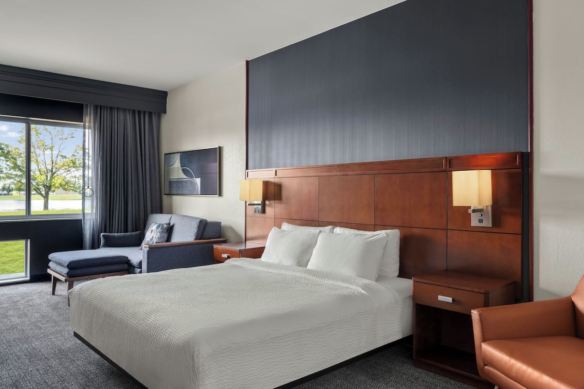 A hotel room with a large bed, a desk, a chair, a window with curtains, and a wall-mounted lamp.