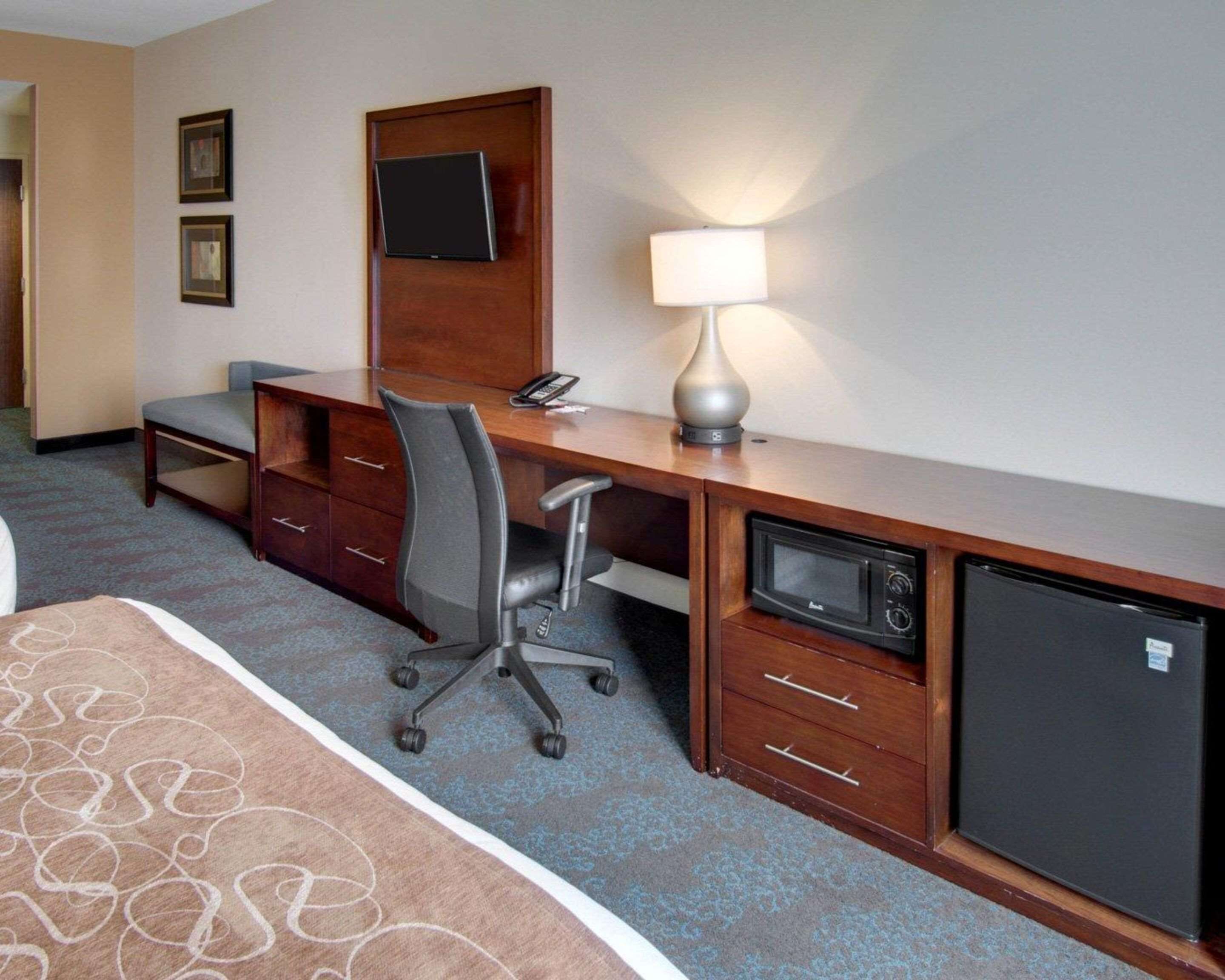 A hotel room with a desk, chair, lamp, microwave, and refrigerator.