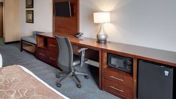 A hotel room with a desk, chair, lamp, microwave, and refrigerator.