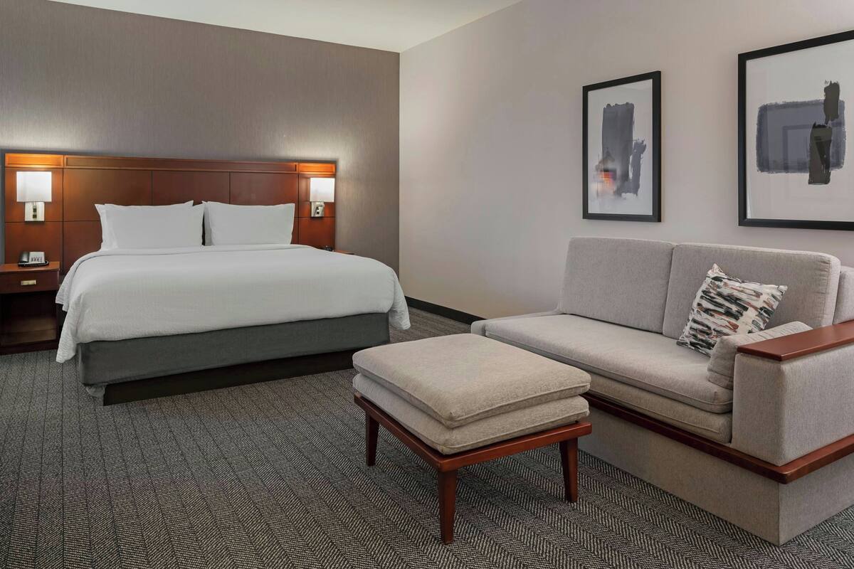 Una habitación de hotel con una cama grande, un sofá, un reposapiés, dos lámparas de pared y dos obras de arte enmarcadas.