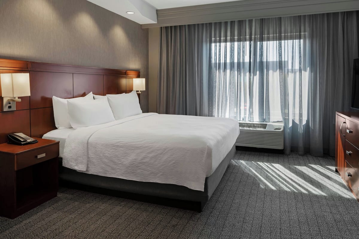 Habitación de hotel con una cama grande, una mesita de noche con teléfono, televisor montado en la pared y una ventana con cortinas.
