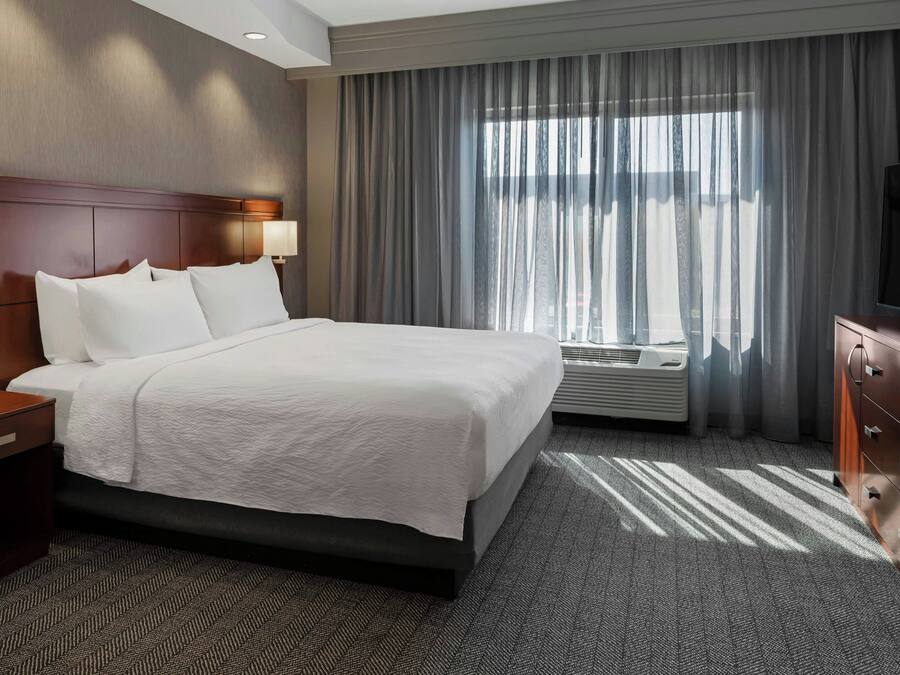 A hotel room with a large bed, a nightstand with a telephone, a wall-mounted TV, and a window with curtains.