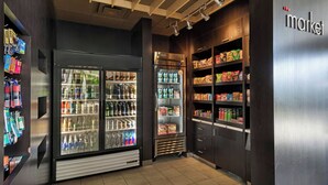 Una kitchenette bien iluminada con un refrigerador, un microondas y una cafetera. Se observan estantes llenos de aperitivos y bebidas.