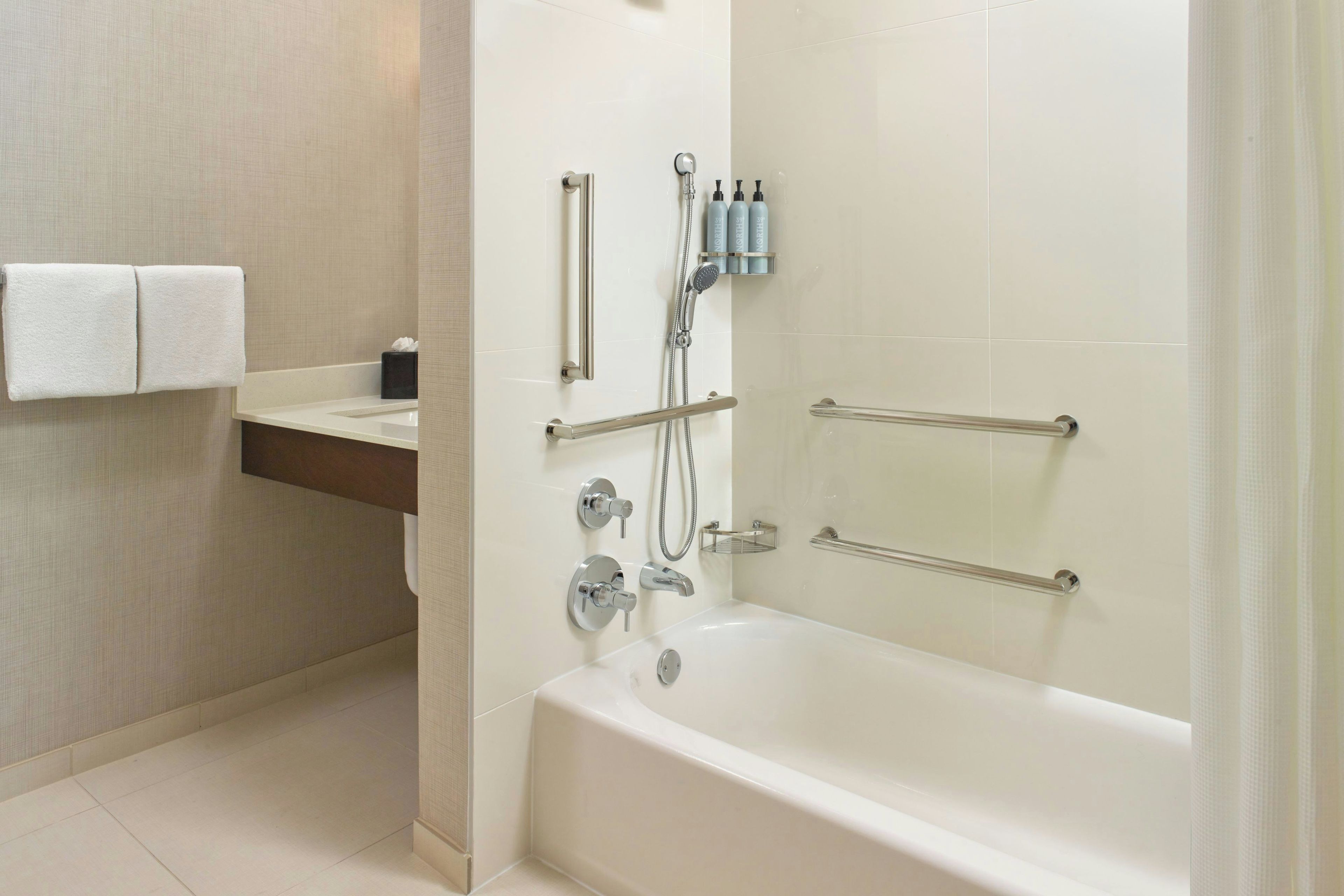 Un baño moderno con bañera, ducha e inodoro. Hay barras de apoyo y un perchero para toallas.