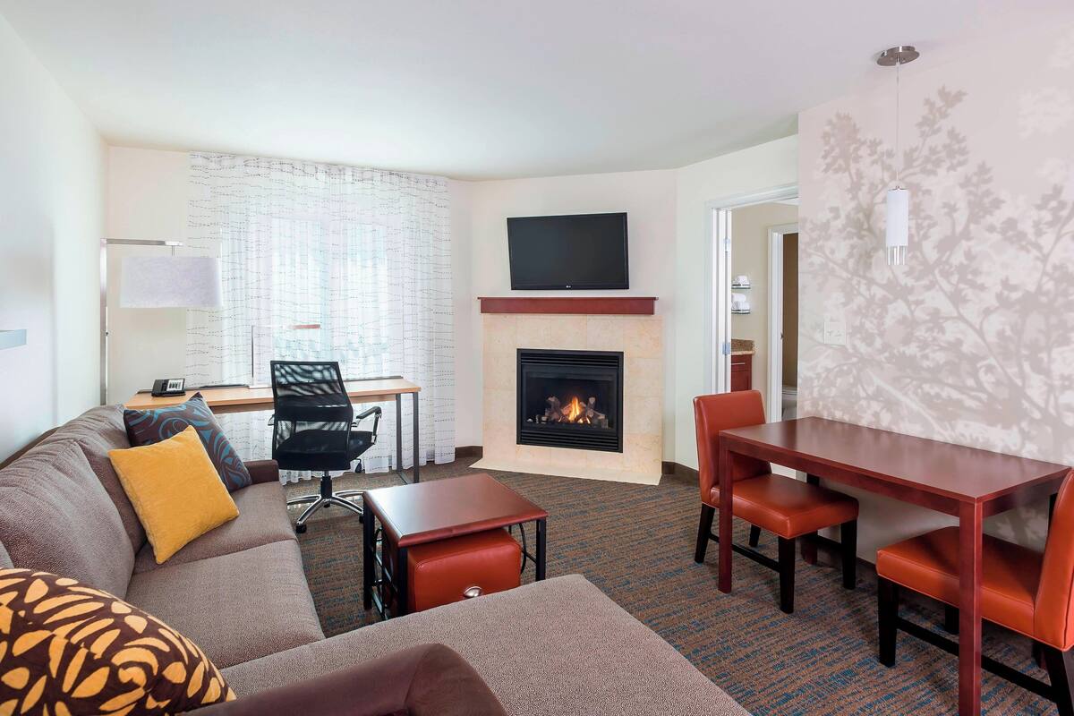 A hotel room with a living area featuring a sofa, a coffee table, a fireplace, a TV mounted above the fireplace, and a dining table with chairs.