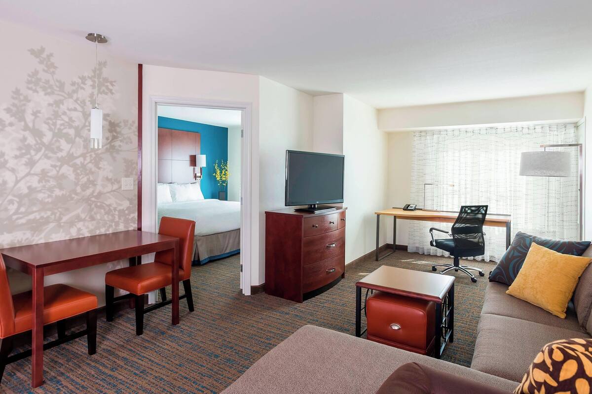 A hotel room with a bed, a desk with a computer, a TV, a dining table with chairs, and a sofa with cushions.