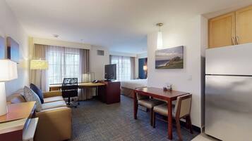 Habitación de hotel con sofá, escritorio, mesa de comedor, televisor, refrigerador y un cuadro en la pared.