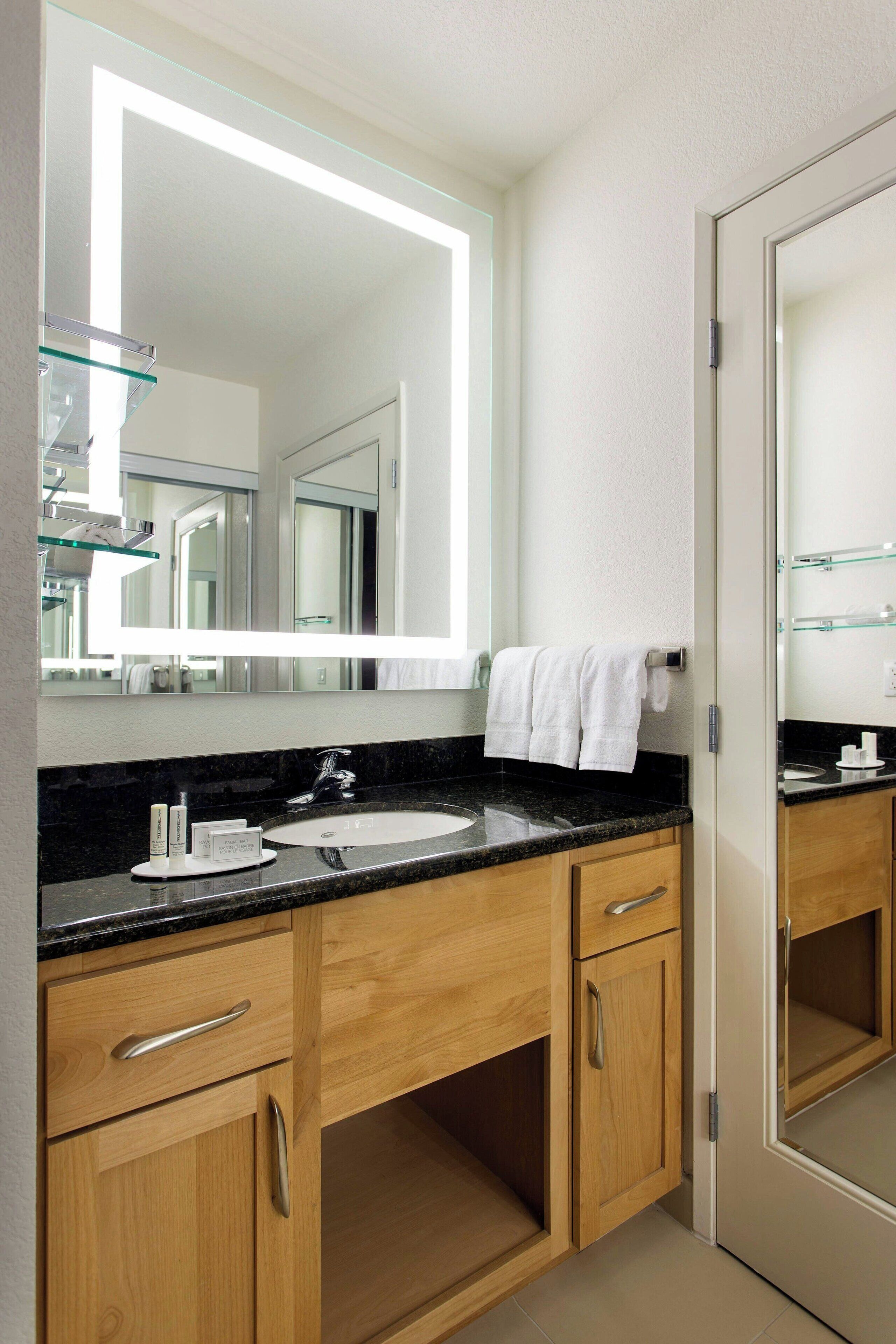 A modern bathroom with a large mirror, a black countertop, wooden cabinets, and a glass shelf.