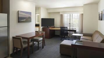Habitación de hotel con zona de comedor, sofá, televisor y ventana con cortinas.