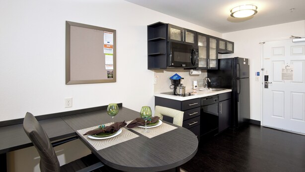 A modern kitchen with dark wood cabinets, a black refrigerator, and a microwave. There is a dining table with two chairs and a placemat. - Candlewood Suites Greeley by IHG (Greeley)