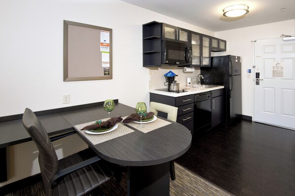 A modern kitchen with dark wood cabinets, a black refrigerator, and a microwave. There is a dining table with two chairs and a placemat. - Candlewood Suites Greeley by IHG (Greeley)