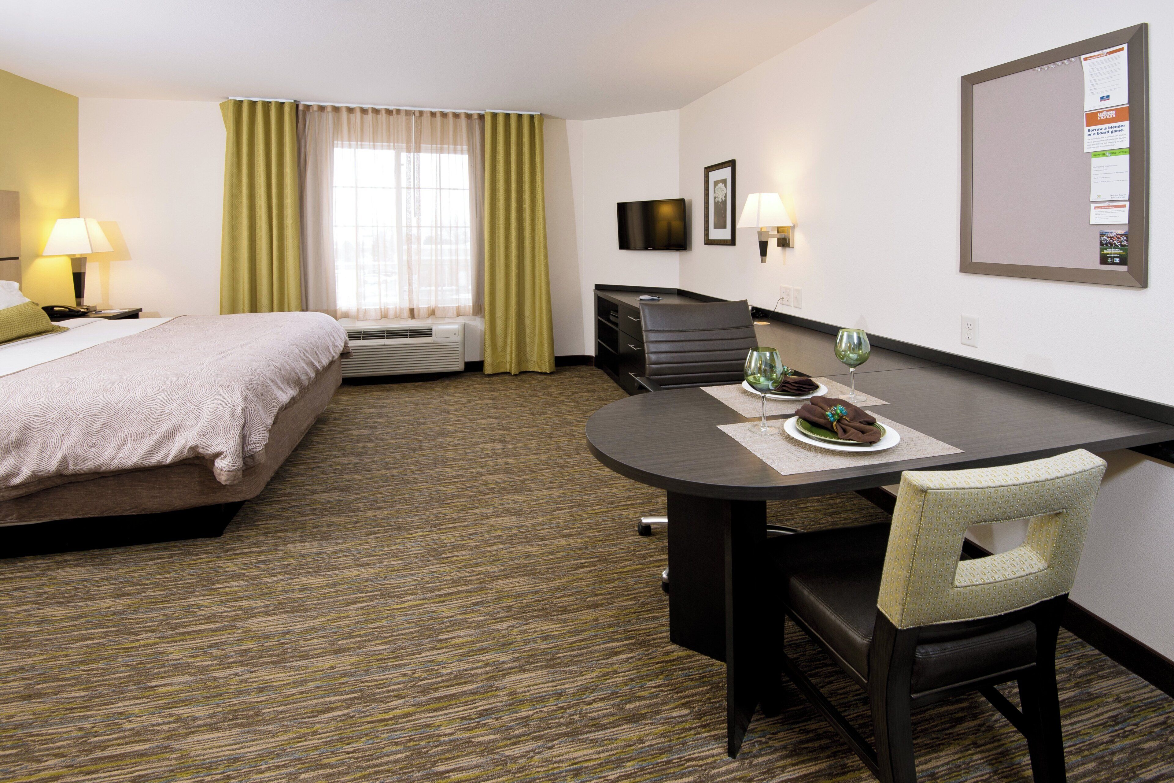A hotel room with a bed, a desk with a chair, a television, and a bulletin board.