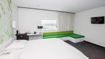 A hotel room with a large bed, a green bench, a desk with a lamp, and a window with curtains.
