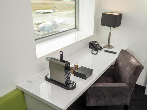 A hotel room with a desk, a coffee machine, a telephone, a remote control, and a window with a view of an airport.