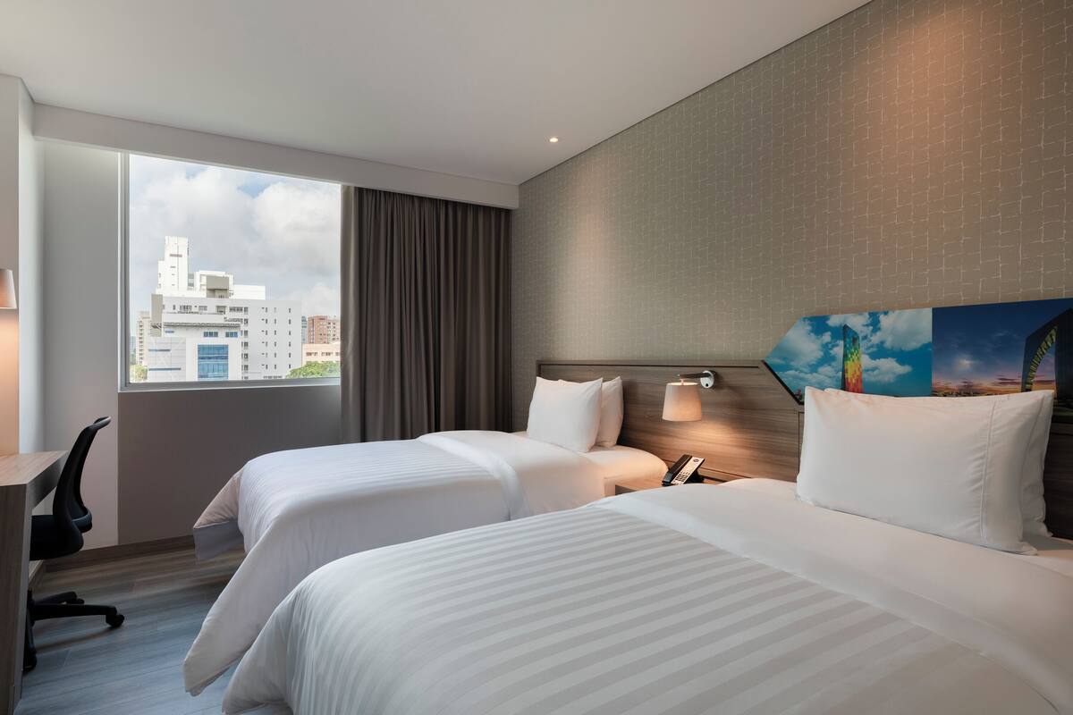 A hotel room with two beds, a desk, a chair, a nightstand, and a city view through the window.