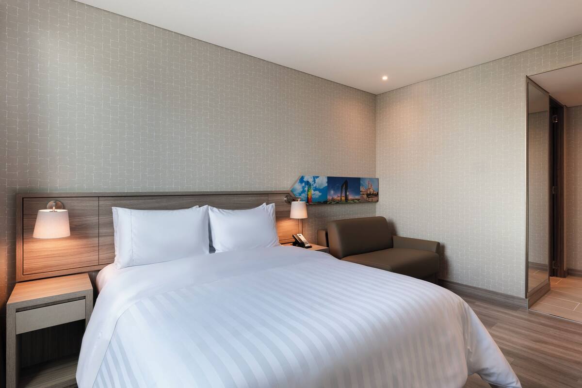 A modern hotel room with a large bed, a nightstand with a lamp, a chair, and a painting on the wall.