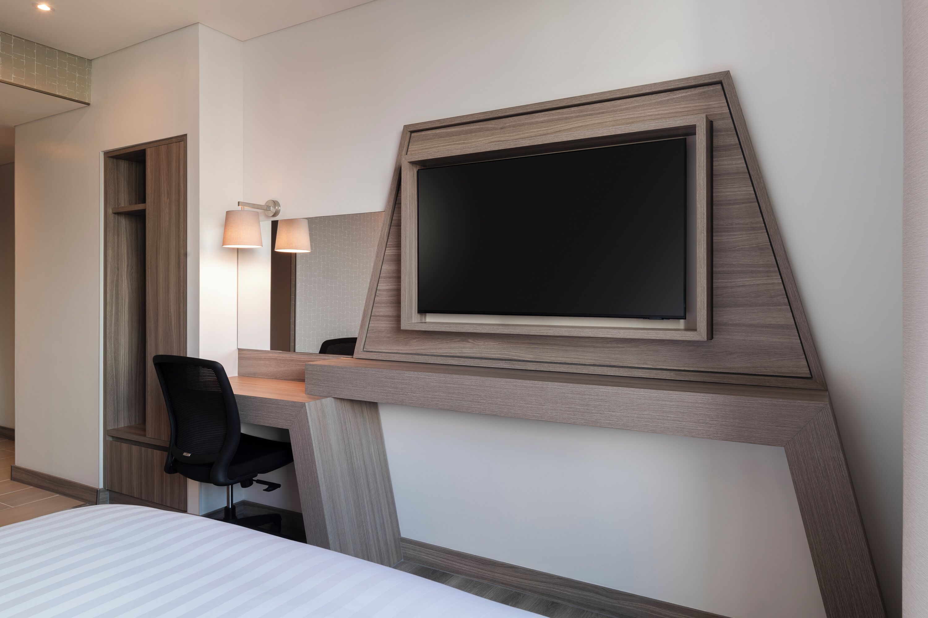 A modern hotel room with a bed, a desk with a chair, a flat-screen TV mounted on the wall, and two bedside lamps.