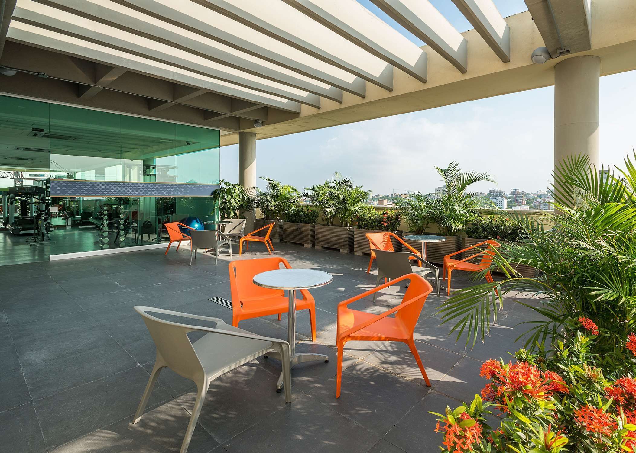 An outdoor patio area with orange chairs and tables, surrounded by greenery and a view of the city skyline.