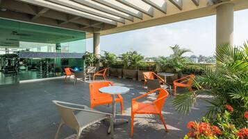 An outdoor patio area with orange chairs and tables, surrounded by greenery and a view of the city skyline.