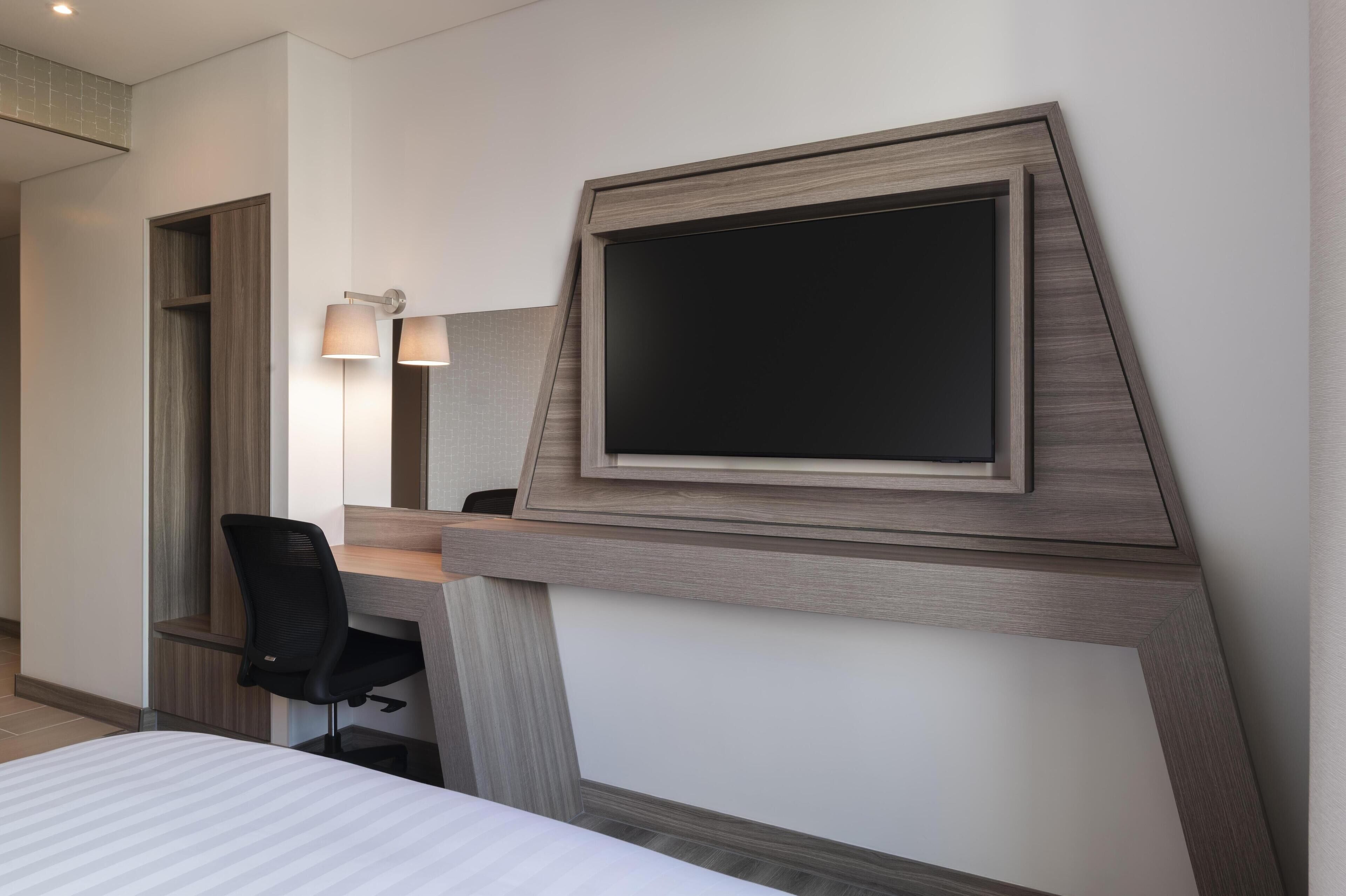 A hotel room with a bed, a desk with a chair, a large mirror, and a flat-screen TV mounted on the wall.
