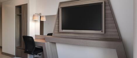 A hotel room with a bed, a desk with a chair, a large mirror, and a flat-screen TV mounted on the wall.