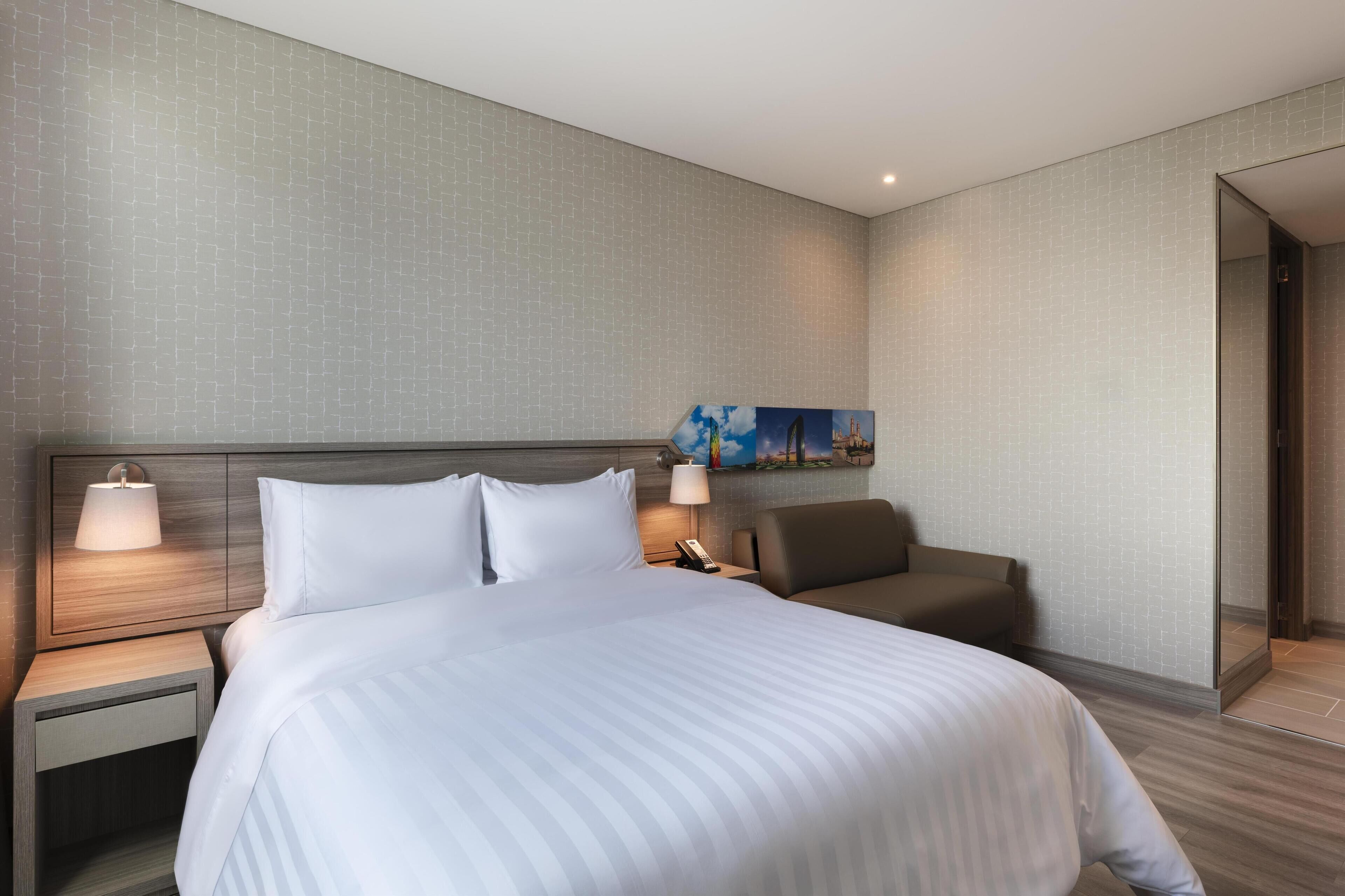 A hotel room with a large bed, a nightstand with a lamp, a seating area with a chair, and a painting on the wall.