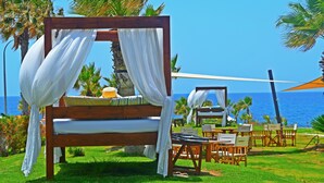 A wooden bed with white drapes, a table with a yellow object, and chairs in a grassy area with a view of the ocean. - Amphora Hotel & Suites (Paphos)