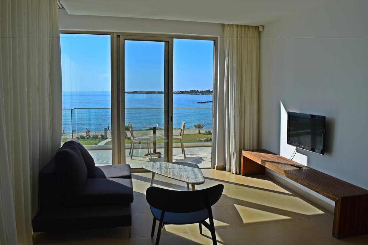 Une chambre moderne avec un canapé, une petite table et un téléviseur. Elle est dotée d’une grande porte-fenêtre coulissante qui donne sur un balcon offrant une vue sur la mer.