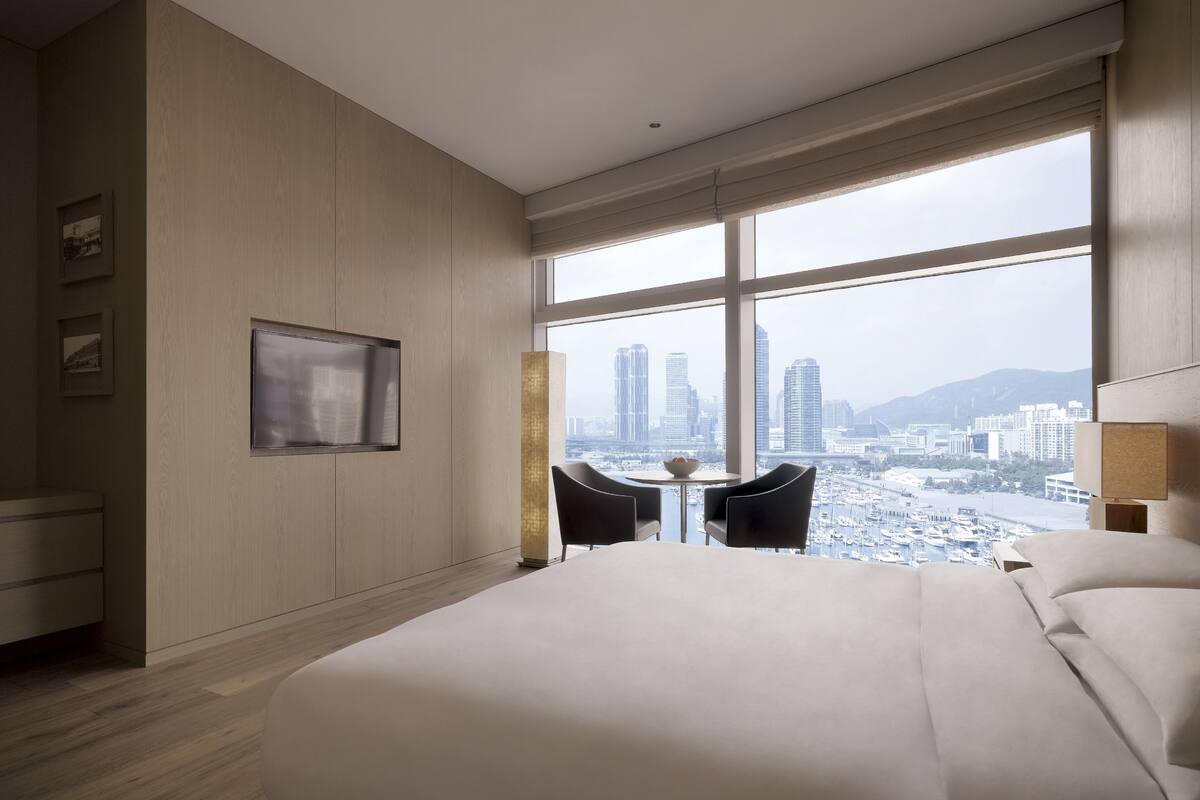 A modern hotel room with a large bed, a small round table, two chairs, a TV, and a city view through large windows.