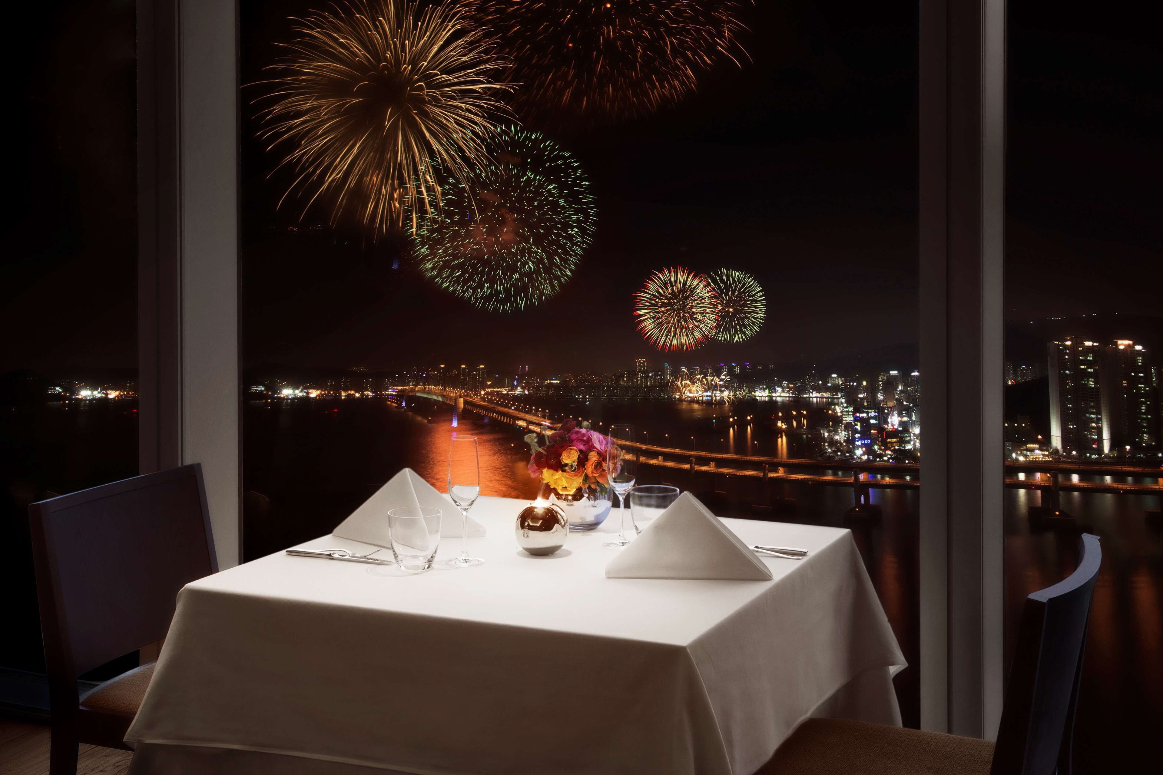 Mesa lista para comer con vista a la ciudad y fuegos artificiales al fondo.