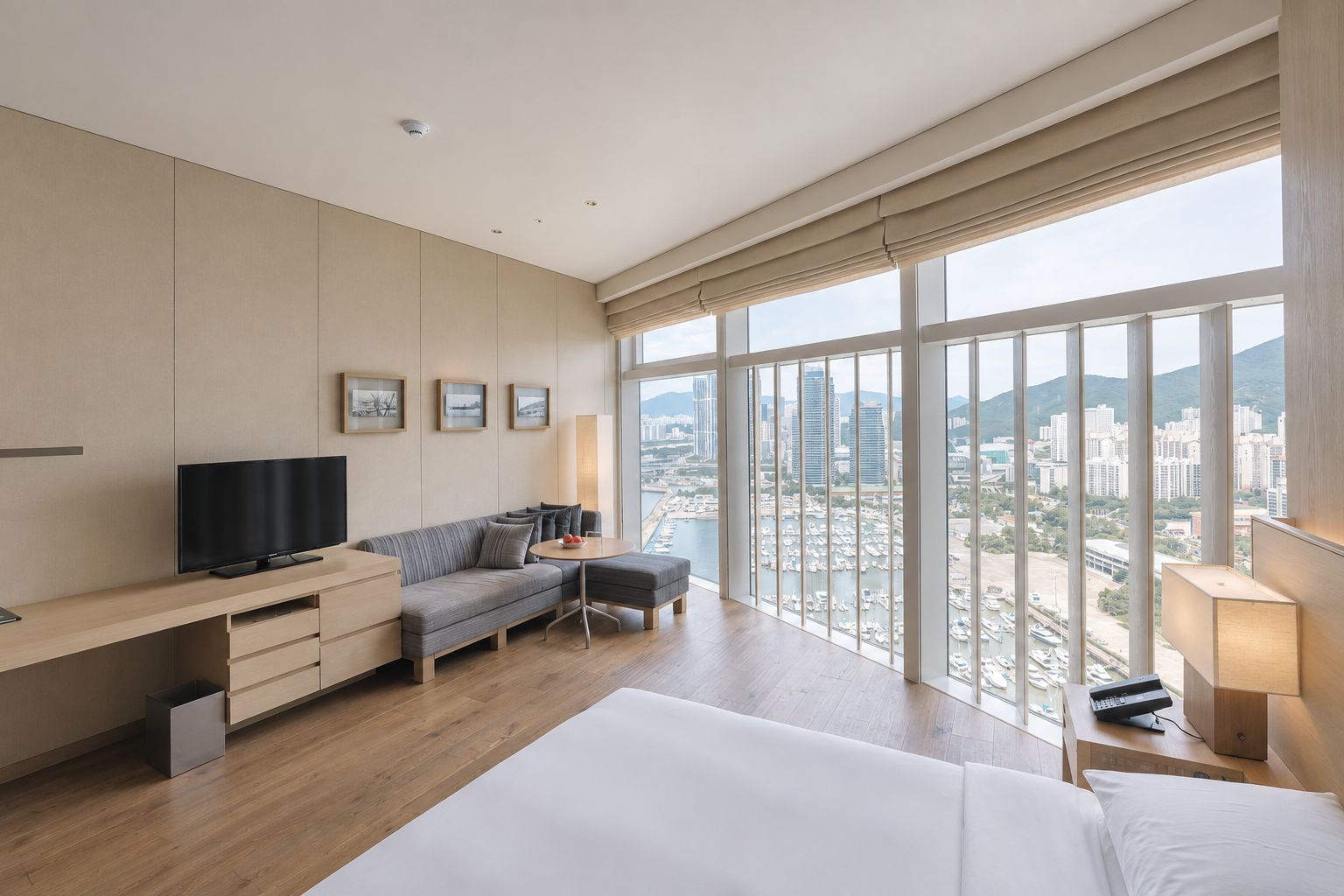 A hotel room with a large bed, a sofa, a desk with a TV, and a view of the city through large windows.