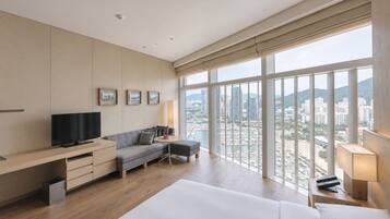 Habitación de hotel con una cama grande, un sofá, un escritorio con televisor y vistas a la ciudad a través de amplios ventanales.