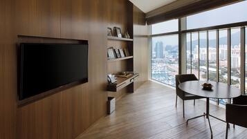 A modern living room with a flat-screen TV, wooden shelving with decorative items, a dining table with chairs, and a large window offering a city view.
