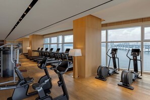 A gym with treadmills, elliptical machines, and weight equipment, large windows with a view of a bridge and cityscape.