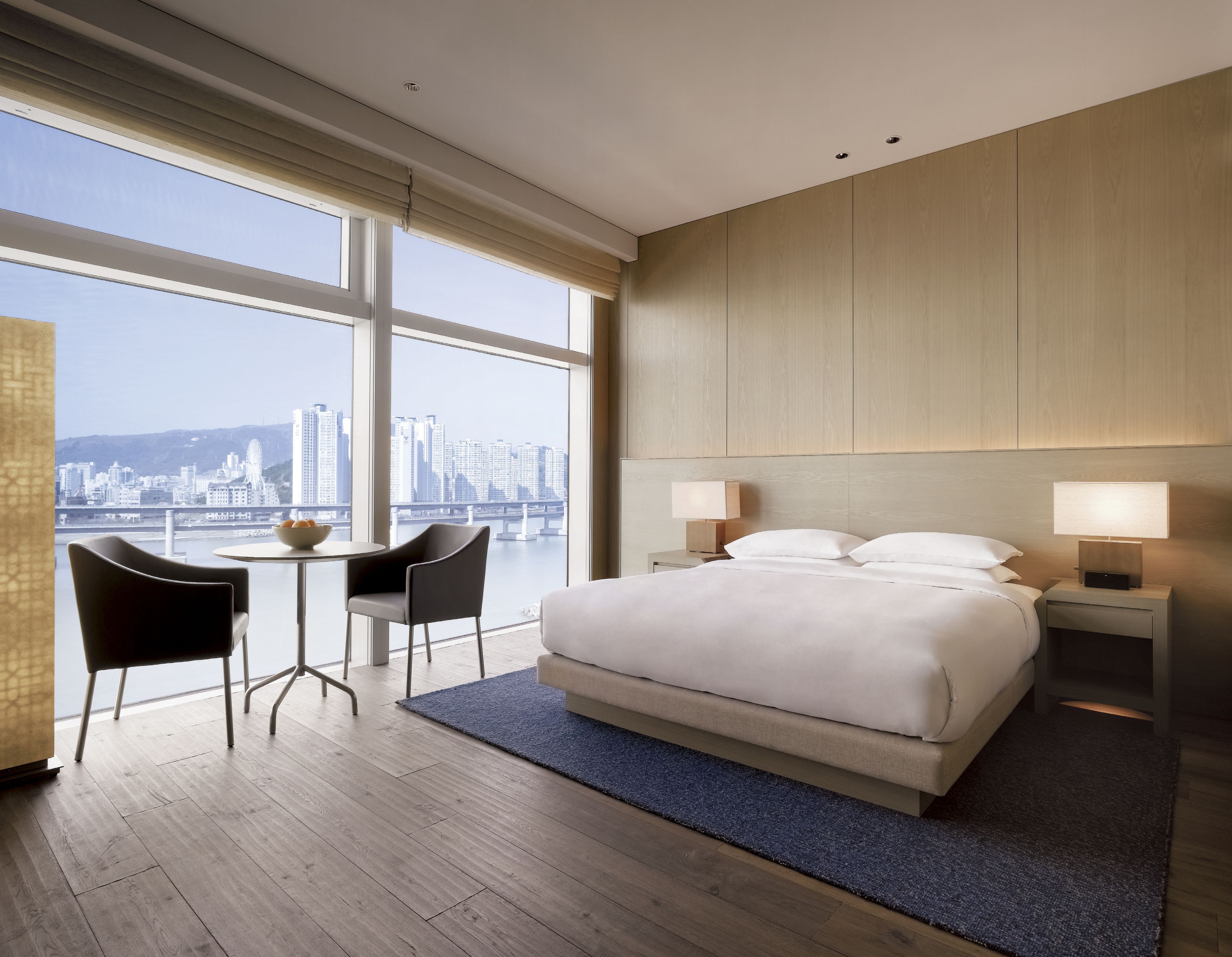 Ein Hotelzimmer mit einem großen Bett, zwei Nachttischen, einem Schreibtisch mit Stuhl und Blick auf eine Stadtlandschaft durch große Fenster.