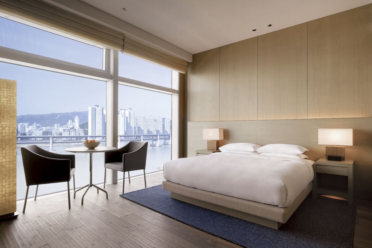 A hotel room with a large bed, two bedside tables, a desk with a chair, and a view of a cityscape through large windows.