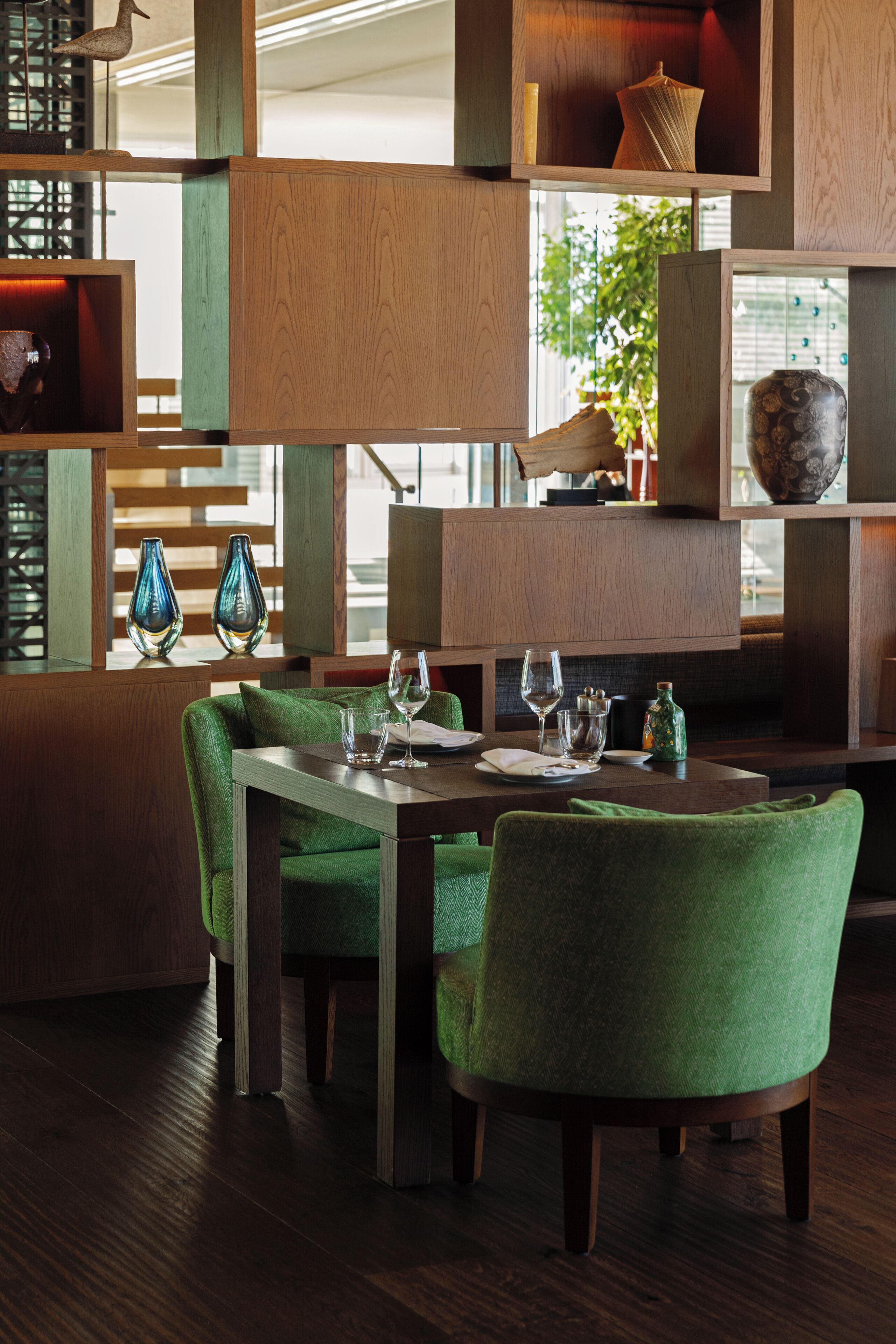 Un comedor con mesa de madera, sillas tapizadas de color verde y objetos decorativos en estantes.