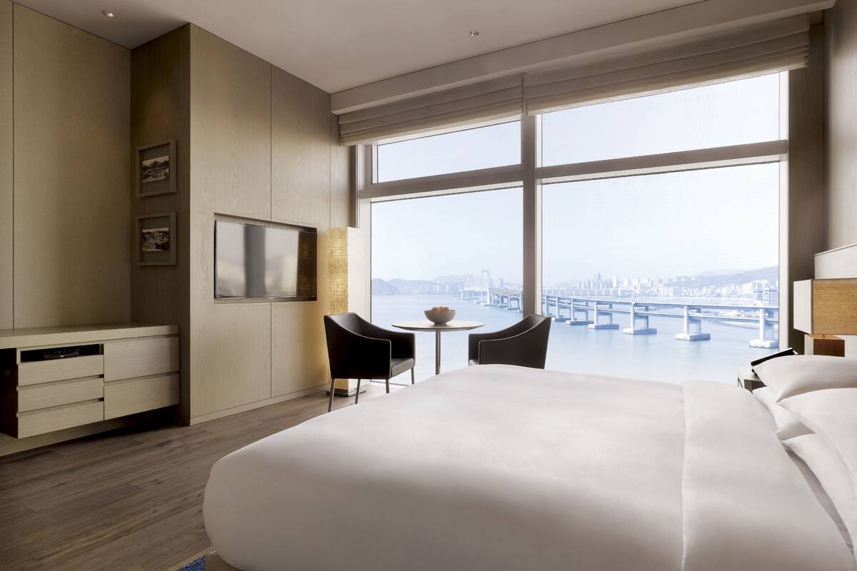 A hotel room with a large bed, a desk, a chair, a small table, and a view of a cityscape and river.