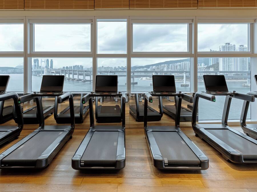 A gym with treadmills and laptops, large windows with a city view.