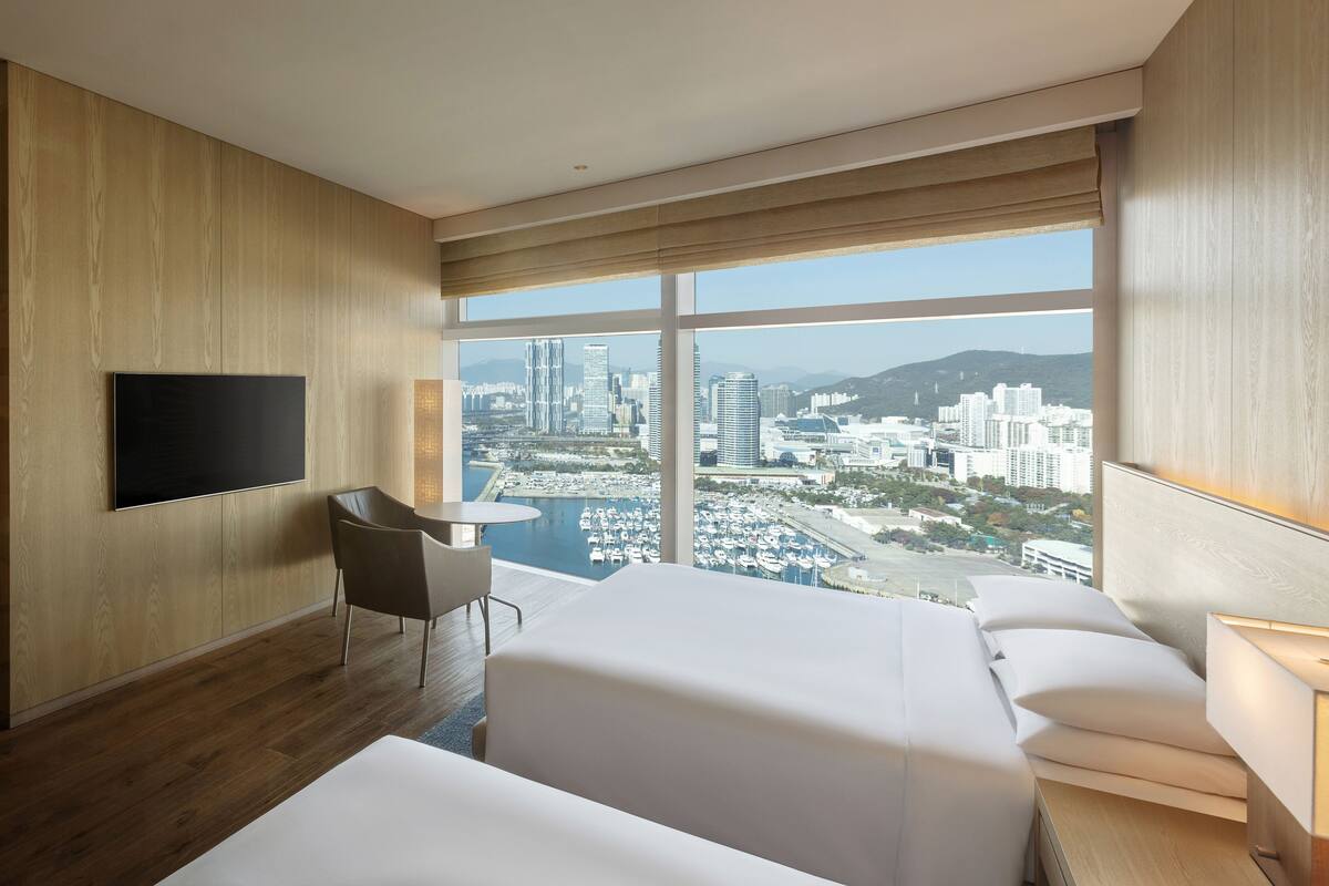 A hotel room with a large window offering a city view, a flat-screen TV mounted on the wall, a small round table, and a sofa.