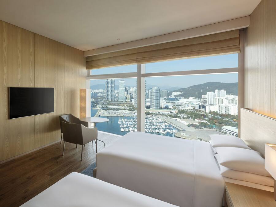 A hotel room with a large window offering a city view, a flat-screen TV mounted on the wall, a small round table, and a sofa.