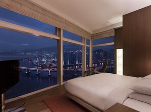 Une chambre d’hôtel moderne avec un grand lit, une télévision et une vue sur le paysage urbain illuminé la nuit.